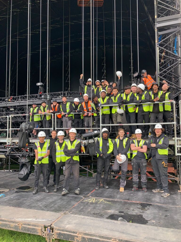 A large group shot of crew wearing high viz and hard hats, stood on a stage at a festival.