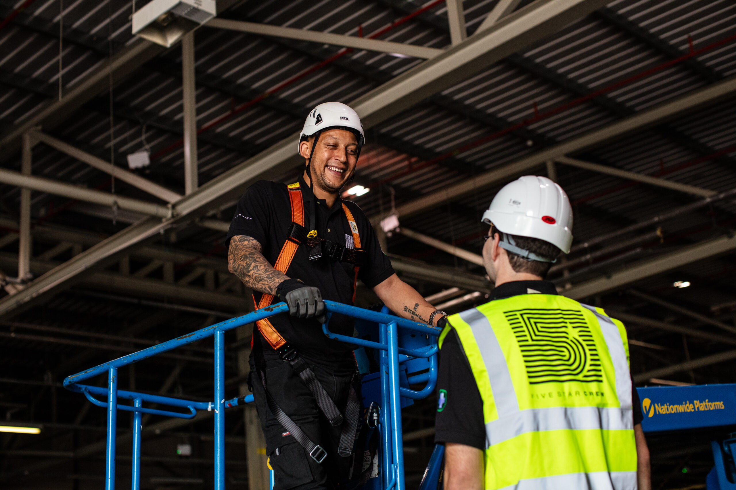 Crew smiling at each other. One is wearing a harness and is fastened onto plant, and the other is wearing a high viz. Both are wearing hharrd hats